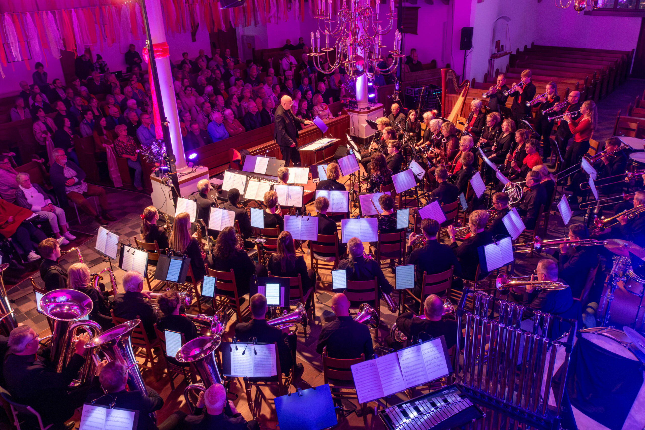 Een groot harmonieorkest treedt op in een sfeervol verlichte kerk met paars en roze licht voor een vol publiek.